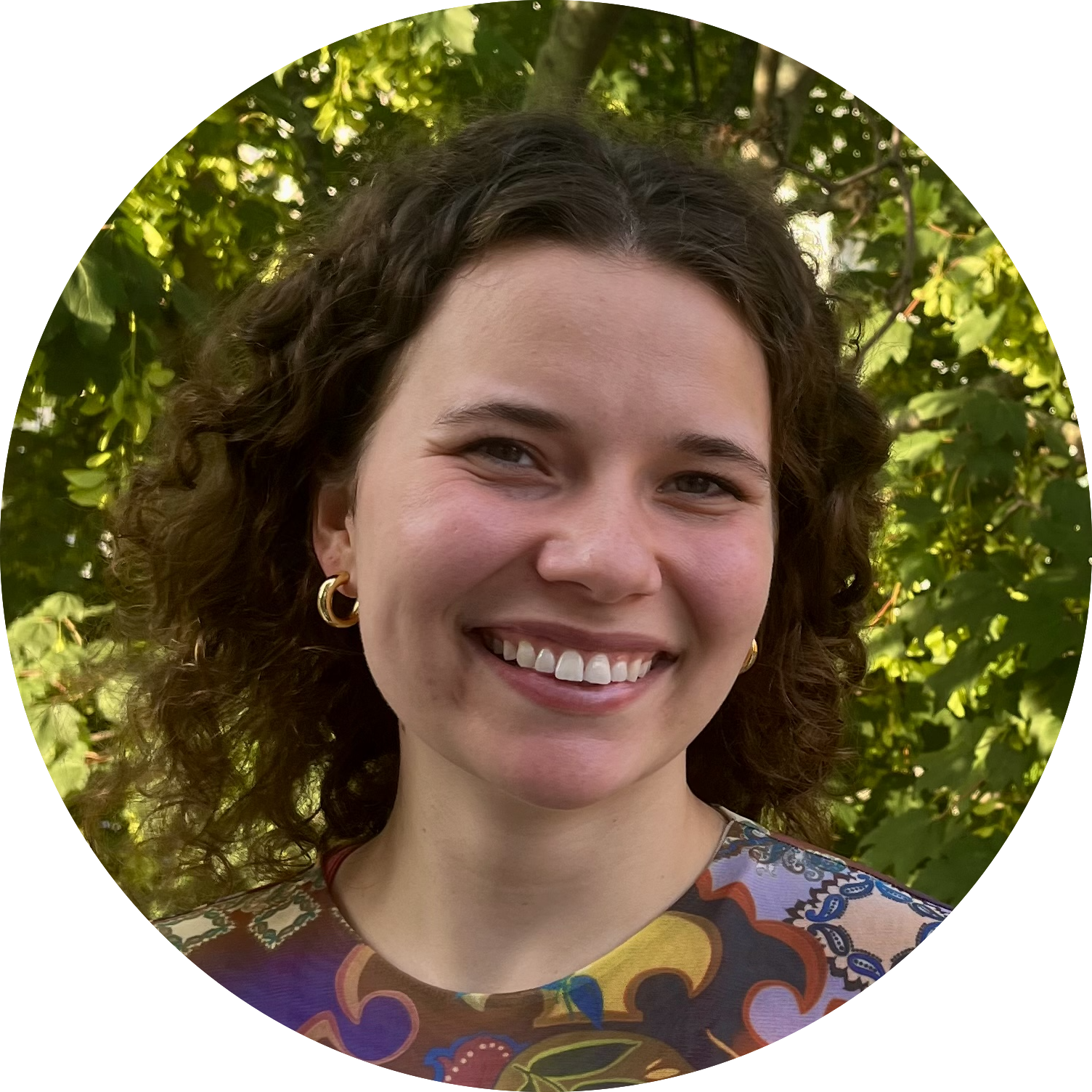 Picture of Margaret Corona - a young woman with olive skin and brown hair with a radiant smile wearing a colorful shirt in front of a tree with many leaves