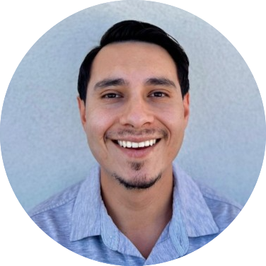 Smiling person with short dark hair and a goatee, wearing a light gray collared shirt, against a light background.