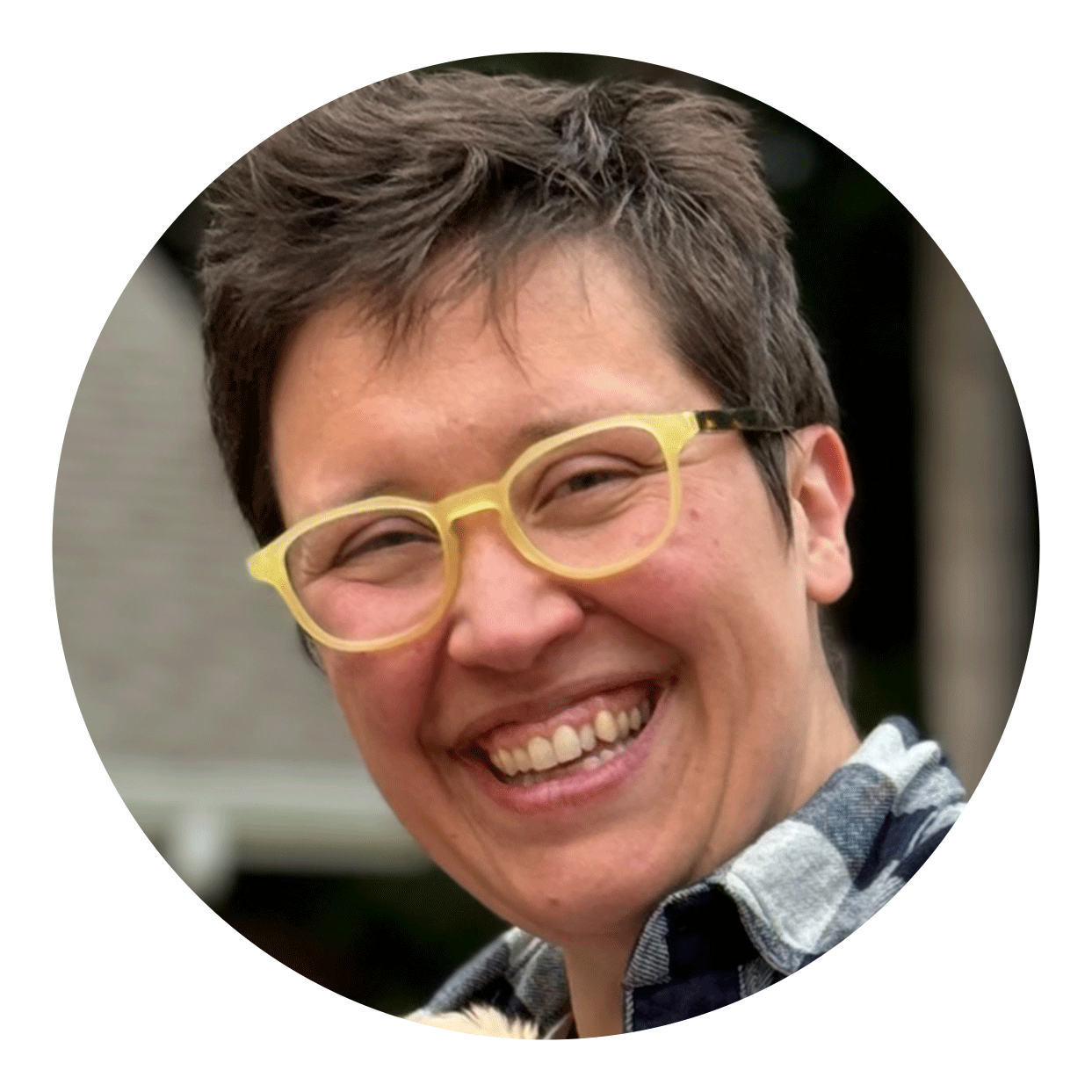 Smiling person with short hair and yellow glasses, wearing a plaid shirt, outdoors.