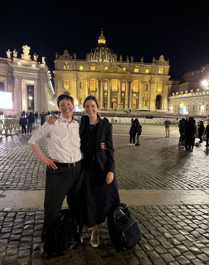 Two people smiling and posing in front of St. Peter's Basilica at night, with bags on the ground.